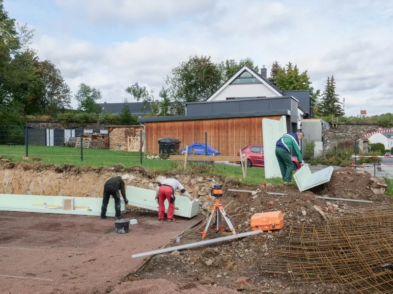 Zwei Arbeiter setzen die gedämmte Randschalung der Bodenplatte, davor der Rotationslaser
