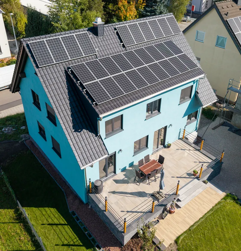 Luftbild des fertigen, hellblauen Wohnhauses mit Photovoltaik, Terrasse und Garten